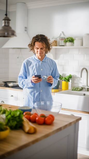 istock Happy Young Man with Long Hair Using Smartphone while Wearing Blue Pajamas. He is Scrolling News Feed, Social Media, Chatting and Drining Morning Coffe in a Kitchen with Healthy Vegetables. 1271303783