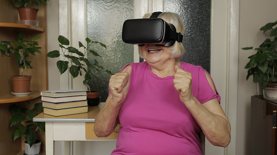 Amazed senior grandmother in virtual headset glasses watching amazing 3d video in 360 vr helmet at home. Future technology. Woman in VR goggles playing games, looking around, shows emotion of surprise