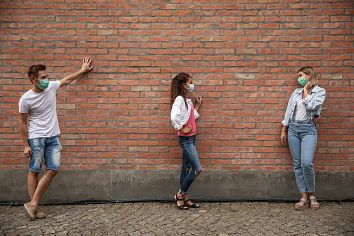 Full length shot of two young women and one young man standing against the wall on the city street and hanging out together. They are keeping social distance and wearing protective face masks.