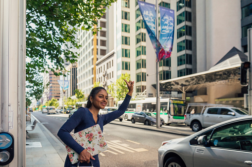 A shot of a businesswoman holding a laptop and handbag, wearing casual business clothing. She is flagging down a taxi in a city on a summers day.