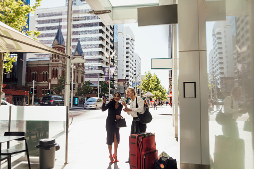 A shot of two business women in a city wearing smart casual business wear. One is standing, holding a mobile phone with luggage and the other is pointing and giving her directions.