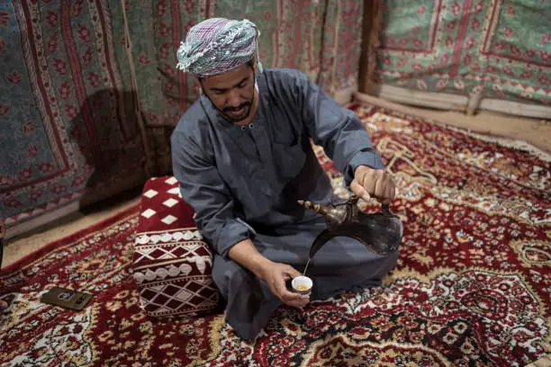 Bedouin man wearing traditional clothes prepares tea on a carpet in the Saudi desert, Saudi Arabia