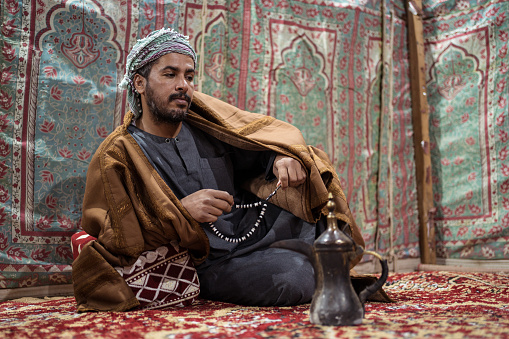 Bedouin man wearing traditional clothes praying with a tasbih while drinking tea on a carpet in the Saudi desert during the night, Saudi Arabia