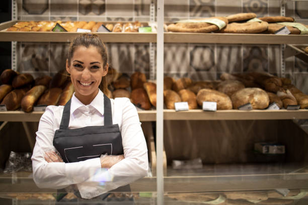 portrait of bakery seller with arms crossed standing in front of shelf full of bred bagels and pastry. - supermarket sales clerk grocer apron imagens e fotografias de stock