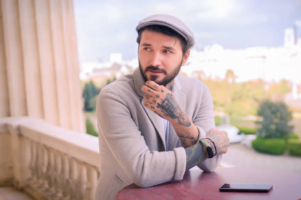 istock Tattooed Man Leaning on a Wooden Table and Playing With His Beard, at the Balcony, While Keeping His Cell Phone On the Table, With City View in the Background 1271312170