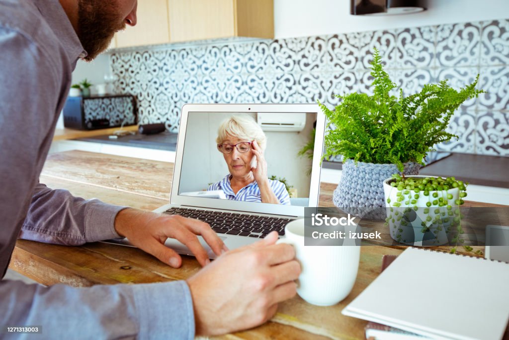 Young man during video call with grandmother Young man having video call with his grandmother, using laptop, sitting at the table in the kitchen at home and drinking coffee. Adult Stock Photo