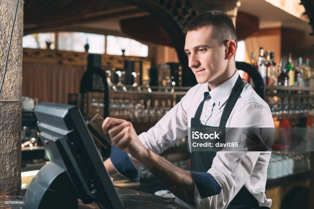small business, people and service concept - happy man or waiter in apron at counter with cashbox working at bar or coffee shop small business, people and service concept - happy man or waiter in apron at counter with cashbox working at bar Point Of Sale Stock Photo