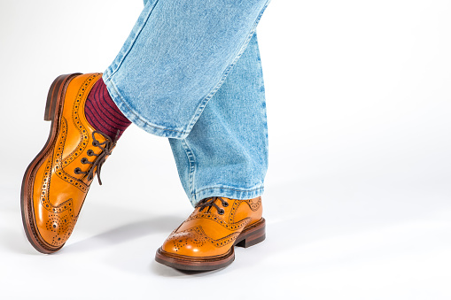 Closeup of Crossed Mens Legs in Brown Oxford Brogue Shoes. Posing in Blue Jeans Against White Background. Horizontal Image