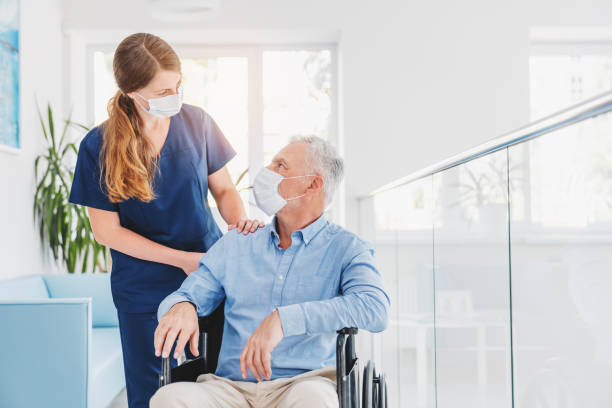 Young woman nurse explaining information to man patient in wheelchair in medical face mask while talking together in hospital. Epidemic and virus concept Young woman nurse explaining information to man patient in wheelchair in medical face mask while talking together in hospital. Epidemic and virus concept medical supplies wheelchair medical equipment nursing home stock pictures, royalty-free photos & images
