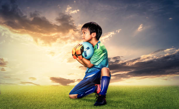 Little Kid goalkeeper holding the ball on a grass field with golden sky background for success soccer dramatic concept. Little Kid goalkeeper holding the ball on a grass field with golden sky background for success soccer dramatic concept. teen goalie stock pictures, royalty-free photos & images