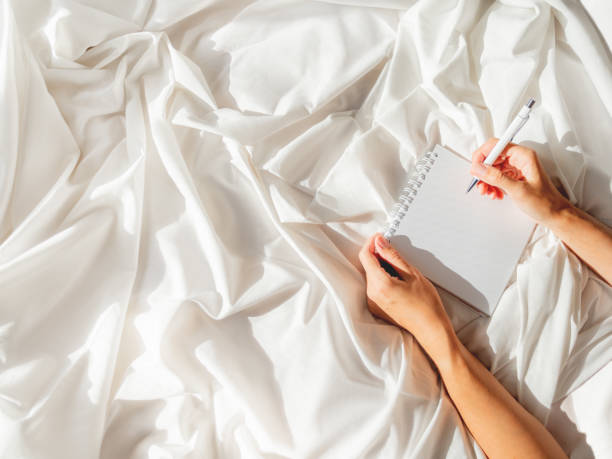 Woman is preparing to write or draw in diary. Top view on crumpled bed sheet and notepad with white paper pages. Practice of positive writing at morning after waking up. Flat lay with copy space.