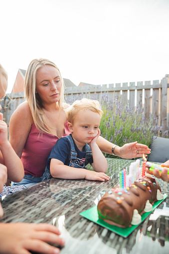 Friends and family celebrating a birthday while sitting outdoors in summer. A mother and son are sitting together at a table while the young boy leans his face on his hand. They are part of the same social bubble who are isolating during the Covid 19 pandemic.