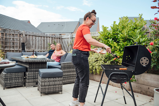 Woman standing outside cooking on a bbq in summer. Her friends are sitting watching