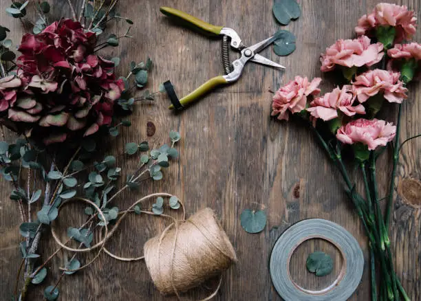 Photo of Florist working space flat lay: purple hydrangea and pink carnation flowers, twine, anchor, pruner and eucalyptus leaves on the rustic wooden table background with space in the middle, top view
