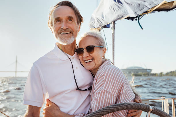 cheerful senior couple standing at steering wheel on a yacht and hugging each other. happy people enjoying sunny day on a boat. - senior adult cheerful adventure discovery imagens e fotografias de stock