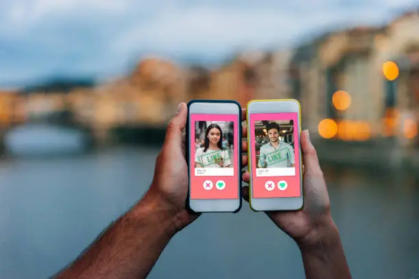Conceptual image of two hands holding smart phones with an online dating app on the screen. Online dating app concept. City at sunset in the background.