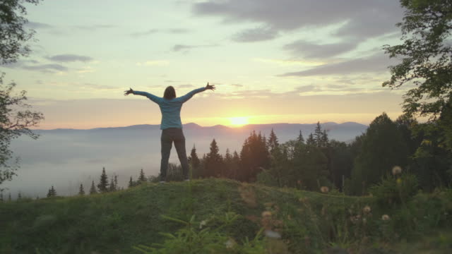 Young brunette woman dancing at the hill and greets the sunrise in mountains. Happy woman enjoys beautiful scenery early in the morning.