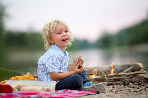 Toddler boy, child having picnic with his family and campfire in the evening near river summertime