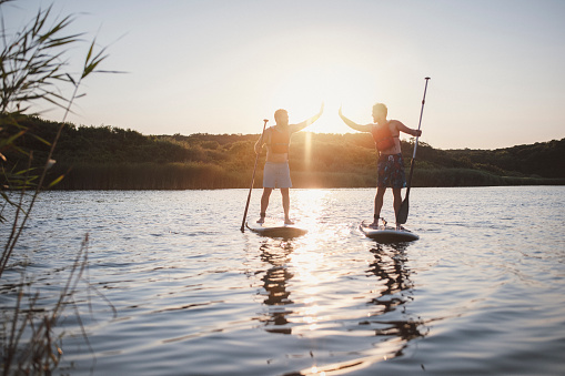 Two young males are paddleboarding at the ocean , giving each other high five , good emotion and summer vibes