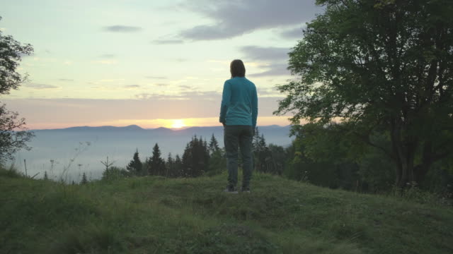 Young female hiker walks on the hill and greets the sunrise in mountains. Happy woman enjoys beautiful scenery early in the morning.