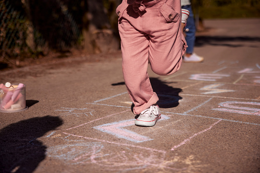 Low section of little girl wearing pink jumpsuit and white shoes jumping hopscotch drawn with chalk outdoors in street
