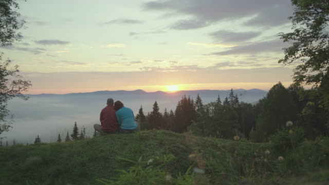 Man and woman sit on the hill and greets the sunrise in mountains. Happy couple enjoys beautiful scenery early in the morning.