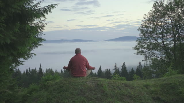 Male hiker walks on the hill and sits to meditate in cloudy mountains. Man enjoys meditation practice in beautiful scenery early in the morning.
