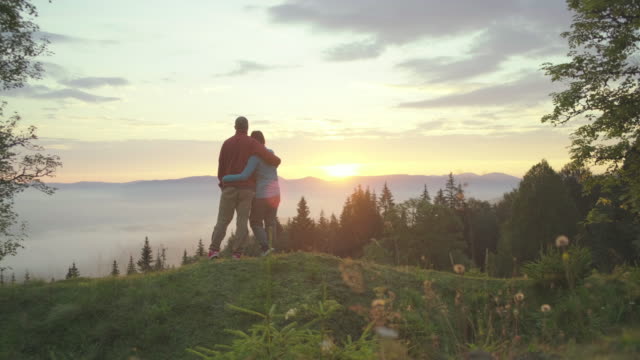 Man and woman walk on the hill and greet the sunrise in mountains. Happy couple enjoys beautiful scenery early in the morning.