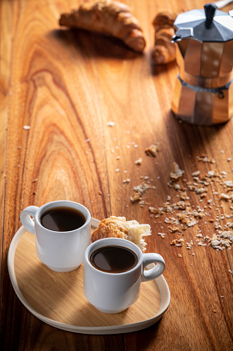 Two coffee cups with espresso black coffee breakfast with croissant on a tray and coffee maker in wooden messy crumbs table