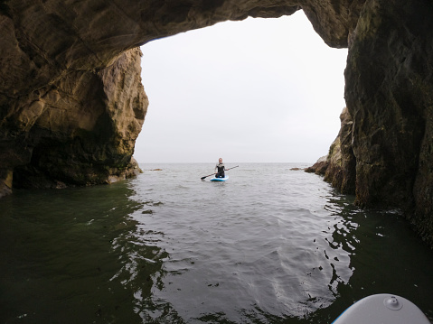 Wide shot of a mid-adult woman paddleboarding through an archway of rocks in the North Sea in Northumberland.