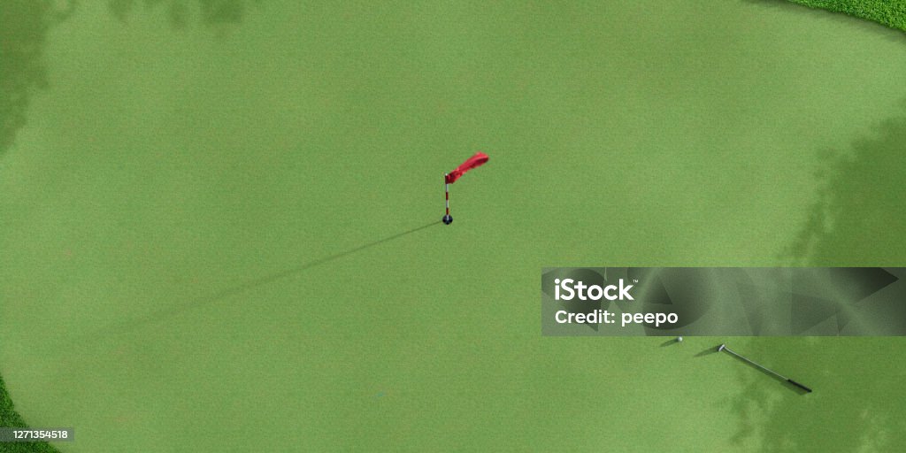 Overhead View of Golf Hole with Flagstick, Putter and Golfball An overhead view of a hole on a golf course with a flagstick with red flag in the middle of the image. The golfball and putter lie near the edge, and the green is framed by longer grass and dappled are shadows. With copy space. Golf Stock Photo