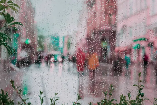 Photo of Abstract blurred background of walking girls, city street after rain, colorful bokeh. View through wet window with raindrops