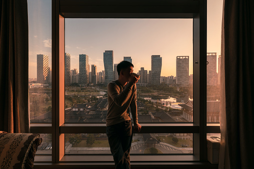 Young asian man standing with drinking coffee in a hotel. Sunrise over modern buildings in Songdo central park at Incheon, South Korea