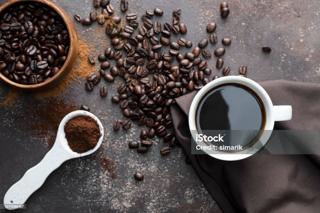 Coffee Direct above view of roasted coffee beans, white coffee cup with black coffee, dark brown colored napkin with one white spoon full with blended coffee on dark brown background. Coffee - Drink Stock Photo