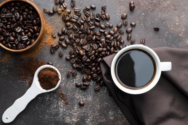 Coffee Direct above view of roasted coffee beans, white coffee cup with black coffee, dark brown colored napkin with one white spoon full with blended coffee on dark brown background. coffee addict stock pictures, royalty-free photos & images