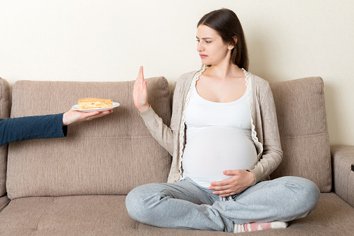 Pregnant woman sitting on the sofa refuses delicious slices of cakes and makes stop gesture. Diet during pregnancy concept.