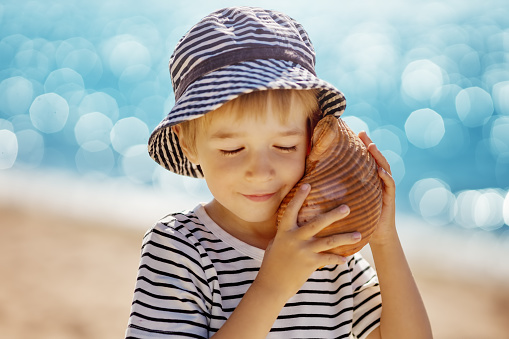 Little boy smiling at the beach in hat and listening to shell.