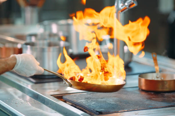 Chef's hands hold a wok with fire over a gas stove. Close-up of the chef's hands cooking food on the fire. Chef prepares food in a professional kitchen.