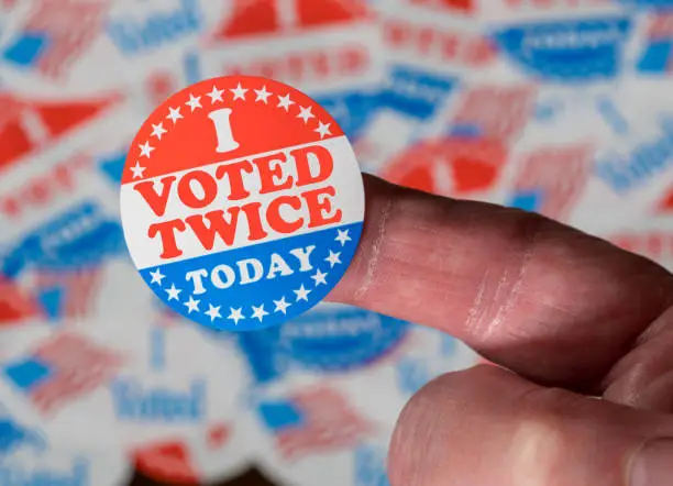 Photo of Finger with I Voted Twice sticker in front of many election voting badges to illustrate voter fraud
