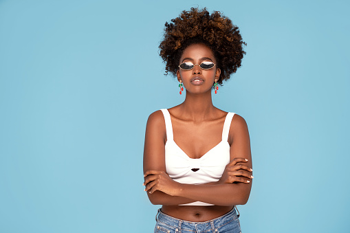 Fashionable young african girl with afro hairstyle posing in studio, wearing sunglasses and earrings. Blue pastel studio background.