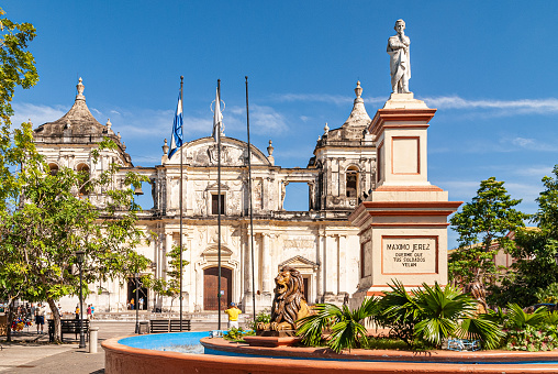 Leon, Nicaragua - November 27, 2008: Combination of statue of Maximo Jerez on beige-maroon pedestal and Cathedral of Assumption in back under blue sky and green foliage in park.