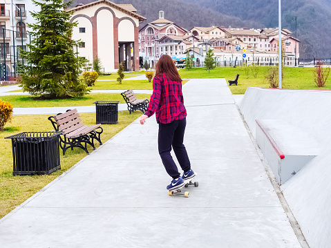 Sochi, Russia - 26 December 2019. A girl in a plaid shirt rides skateboard on a concrete sidewalk in a resort town among the forest and mountains