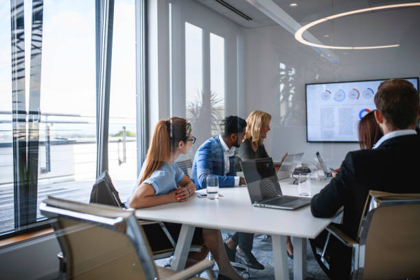 Executive Team Watching Video in Office Conference Room Rear reflective viewpoint of male and female executive team sitting in conference room and watching video. corporate boardroom stock pictures, royalty-free photos & images