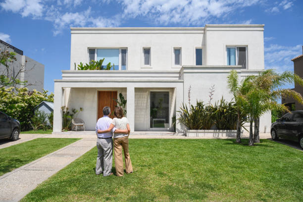 Rear view of senior couple in 60s and 70s standing in front yard with arms around each other and admiring two-story Buenos Aires family home.