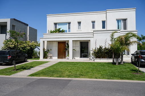 Wide angle street view of modern two-story home in Buenos Aires with well-tended front yard, pedestrian walkway leading to entrance, and cars in driveways.