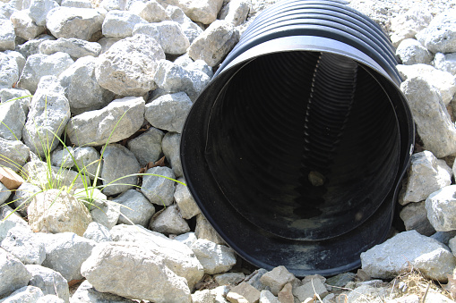 Black plastic pipe for storm water coming out of the ground. Water inside pipe reflecting what's on the other side of the tunnel - surrounded by large rocks. Room for copy on left