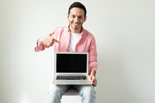 Good looking Hispanic young man sitting on a chair and pointing at a laptop screen with a smile