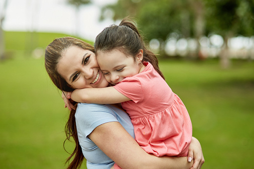 Close-up of 38 year old woman holding 4 year old daughter in her arms and smiling at camera as they spend leisure time together in Miami public park.