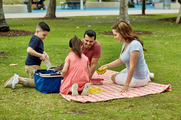 Young Family Unpacking Picnic Basket for Lunch at Miami Park Hispanic mother and father with pre-adolescent children sitting on blanket at park and unpacking sandwiches, fruit, and water for picnic lunch. picnic blanket stock pictures, royalty-free photos & images