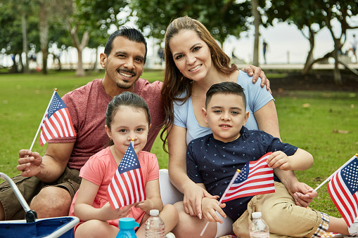 Front view of mid adult mother and father sitting with pre-adolescent children enjoying picnic lunch and holding small American flags as they smile at camera.